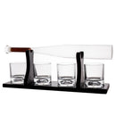 A whiskey decanter set with a clear glass bottle resting horizontally on a black metal stand, with four matching clear glass tumblers on the base, photographed on a white background.