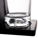 Clear glass tumbler with a thick base resting on a black square coaster or stand, photographed against a white background.