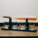 A whiskey decanter and four matching rocks glasses arranged on a black tray, with the decanter tilted horizontally to show the amber liquid inside. The decanter has a clear glass body and a brown wooden stopper, set on a tabletop against a plain light background.