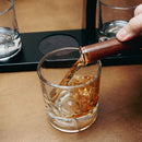 A clear cut-glass rocks tumbler with a textured pattern, being filled with amber whiskey from a dark wooden pouring vessel held by a hand, set on a warm brown tabletop.