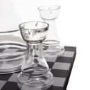 Clear glass chess-piece-shaped candle holders arranged on a black-and-white checkered surface against a white background.