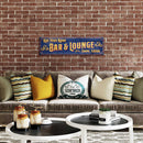 Decorative sign for a bar and lounge above a couch with pillows against a brick wall.