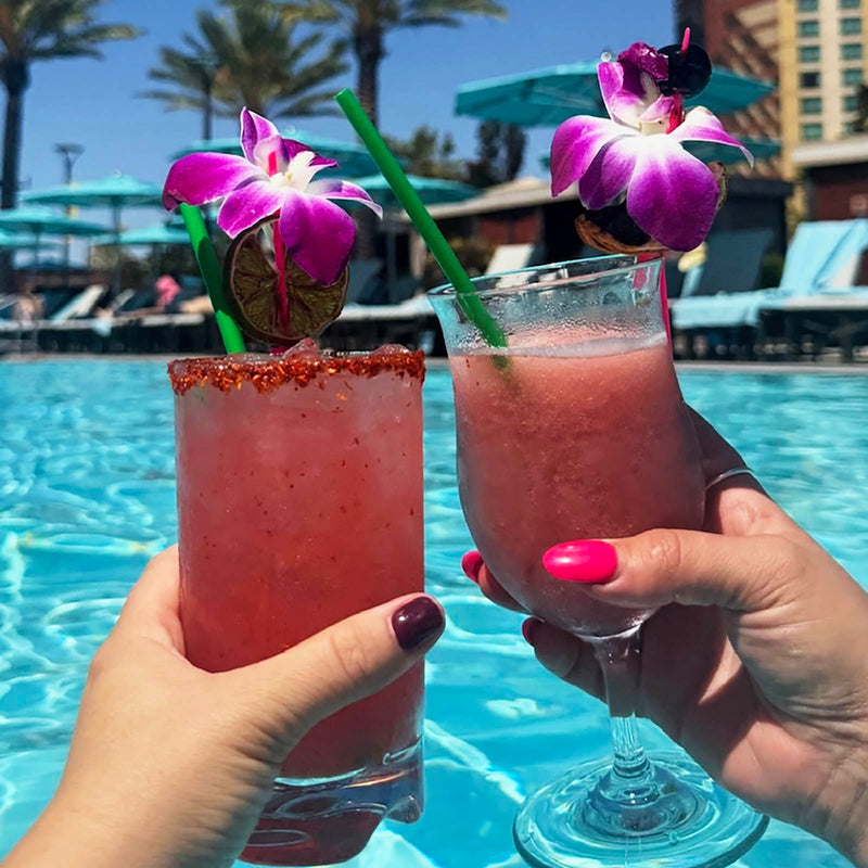 Two pink cocktails with floral garnishes held by hands against a poolside background.