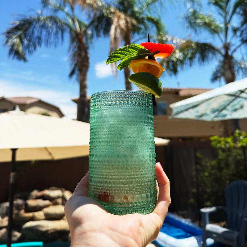 glass with a cocktail garnished with fruit and mint leaves, held in front of a poolside background.