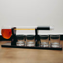 A whiskey decanter set on a black base, featuring a clear glass horizontal tube with a black cap and a glass pitcher filled with amber liquid on the left, plus four small clear glasses on the base.