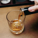 A hand pouring amber-colored liquor from a black-topped glass decanter into a clear cut-glass rocks tumbler on a wooden table.