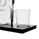 A clear glass tumbler cup sitting on a black rectangular base, with part of a transparent glass decanter visible at the left edge against a white background.
