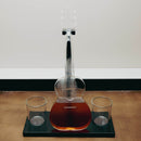 A clear glass decanter shaped like a guitar, filled with amber liquid, displayed on a black stand with two clear glasses on a wooden surface against a plain wall.