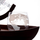 A clear stemless wine glass with an etched world map design, resting on a dark brown wooden tray. A glass decanter is partially visible in the background against a white backdrop.