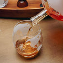 A clear stemless glass with an etched world map design is shown on a wooden surface, with amber-colored liquid being poured from a glass bottle into it.