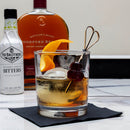 Glass of whiskey cocktail with garnishes on a table with bottles of Woodford Reserve and Fee Brother's Bitters in the background.