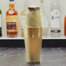 A gold-colored metal cocktail shaker with a textured, dimpled surface and a matching lid, standing on a dark marble countertop with blurred liquor bottles in the background.