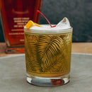 Cocktail in a glass with leaf pattern, garnished with a cherry and orange peel, against a dark background.