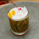 Cocktail with a cherry and orange slice in a glass with leaf patterns on a textured surface.